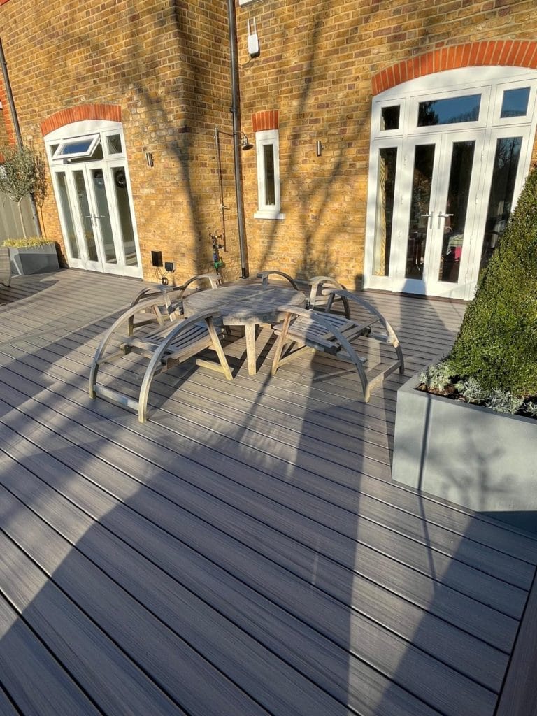 Esher garden with grey composite decking and bespoke curved timber benches beside a brick house, by Flourish Landscaping