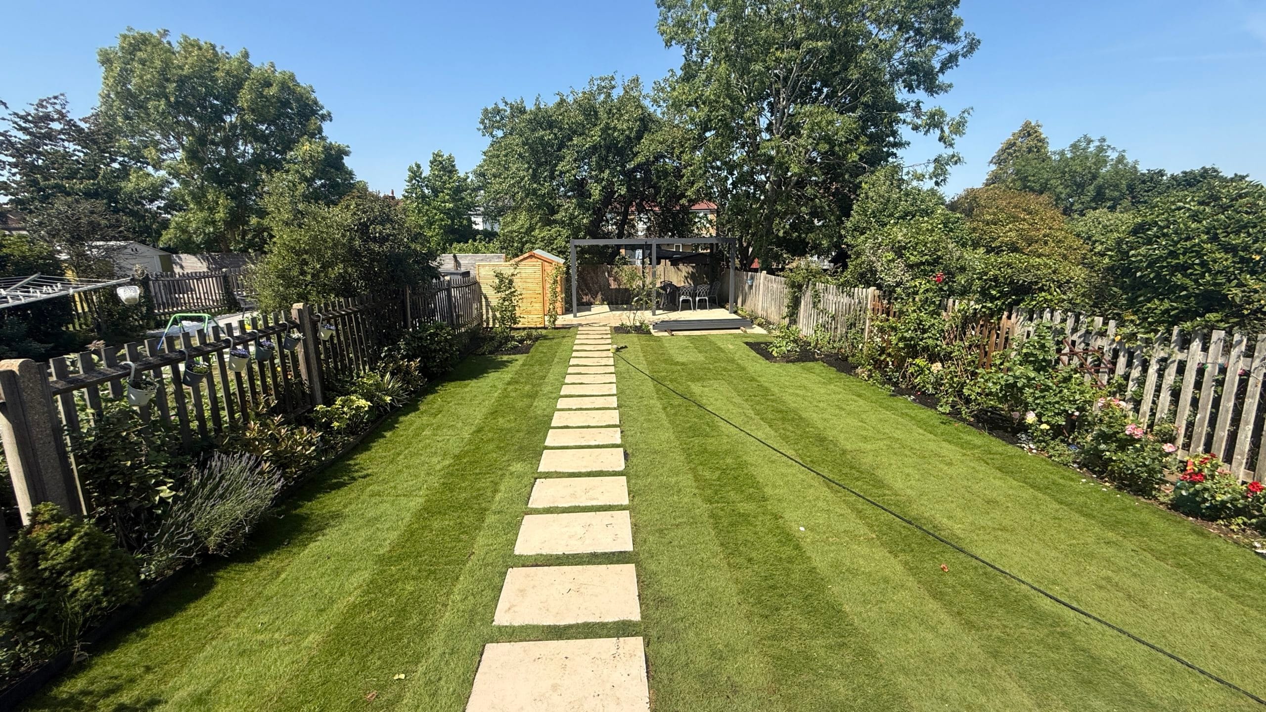 Long view of a large family garden with a striped lawn, central stepping-stone path, mixed borders and mature trees, by Flourish Landscaping