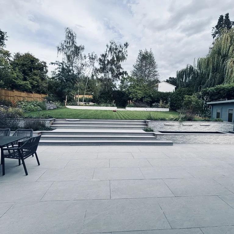 Grey paved terrace with two flights of wide steps, stone-clad retaining walls and a water feature leading up to a striped lawn in a large Surbiton garden by Flourish Landscaping