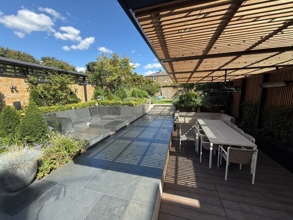A long dining table on a terrace in dappled shade under a slatted timber canopy, beside stone-clad raised beds